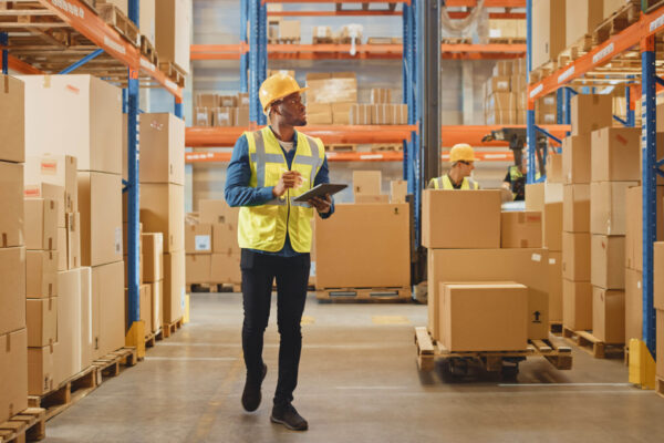 Handsome Male Worker Wearing Hard Hat Holding Digital Tablet Computer Walking Through Retail Warehouse full of Shelves with Goods. Working in Logistics and Distribution Center.