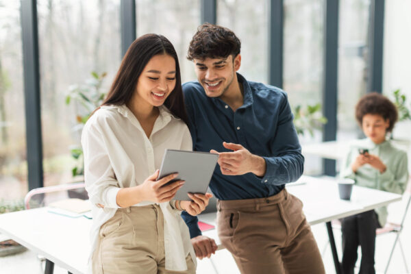 Cheerful Mixed Business Coworkers Couple Using Tablet Computer In Office Digital Fun At Workplace. Cheerful Mixed Business Coworkers Couple Using Tablet Computer Together Surfing Internet And Discussing Corporate News In Modern Office. Teamwork And Interaction