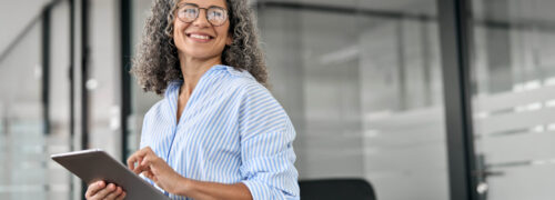 Smiling mature professional business woman bank manager, older happy female executive or lady entrepreneur holding digital tablet pad standing in office at work, looking away at copy space.