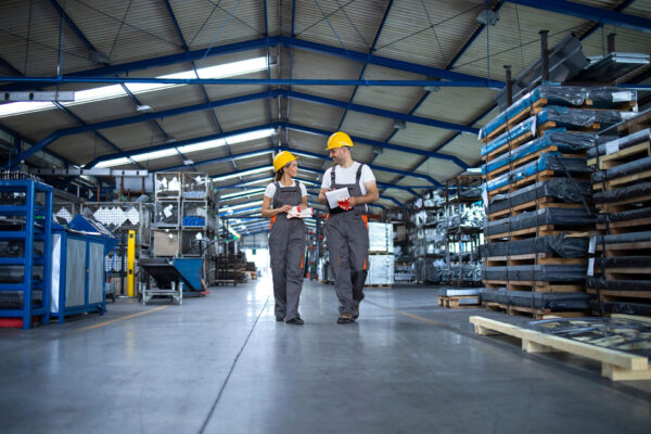 Factory-workers-in-work-wear-and-yellow-helmets-walking-through-industrial-production-hall-and-discussing.