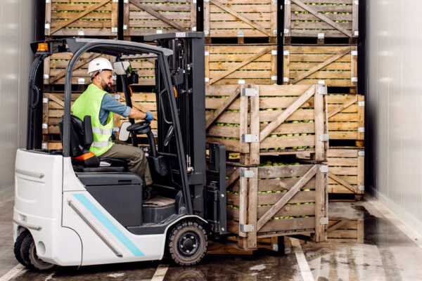 Modern-warehouse,-work-on-forklift,-industry-and-organic-agribusiness.-Smiling-millennial-man-in-helmet-in-forklift-lifts-up-green-apples-in-container-on-shelves-with-many-wooden-crates Modern-warehouse,-work-on-forklift,-industry-and-organic-agribusiness.-Smiling-millennial-man-in-helmet-in-forklift-lifts-up-green-apples-in-container-on-shelves-with-many-wooden-crates