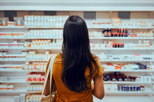 Pharma-Rear-view-of-young-woman-with-bag-standing-against-shelf-in-pharmacy-searching-for-medicine